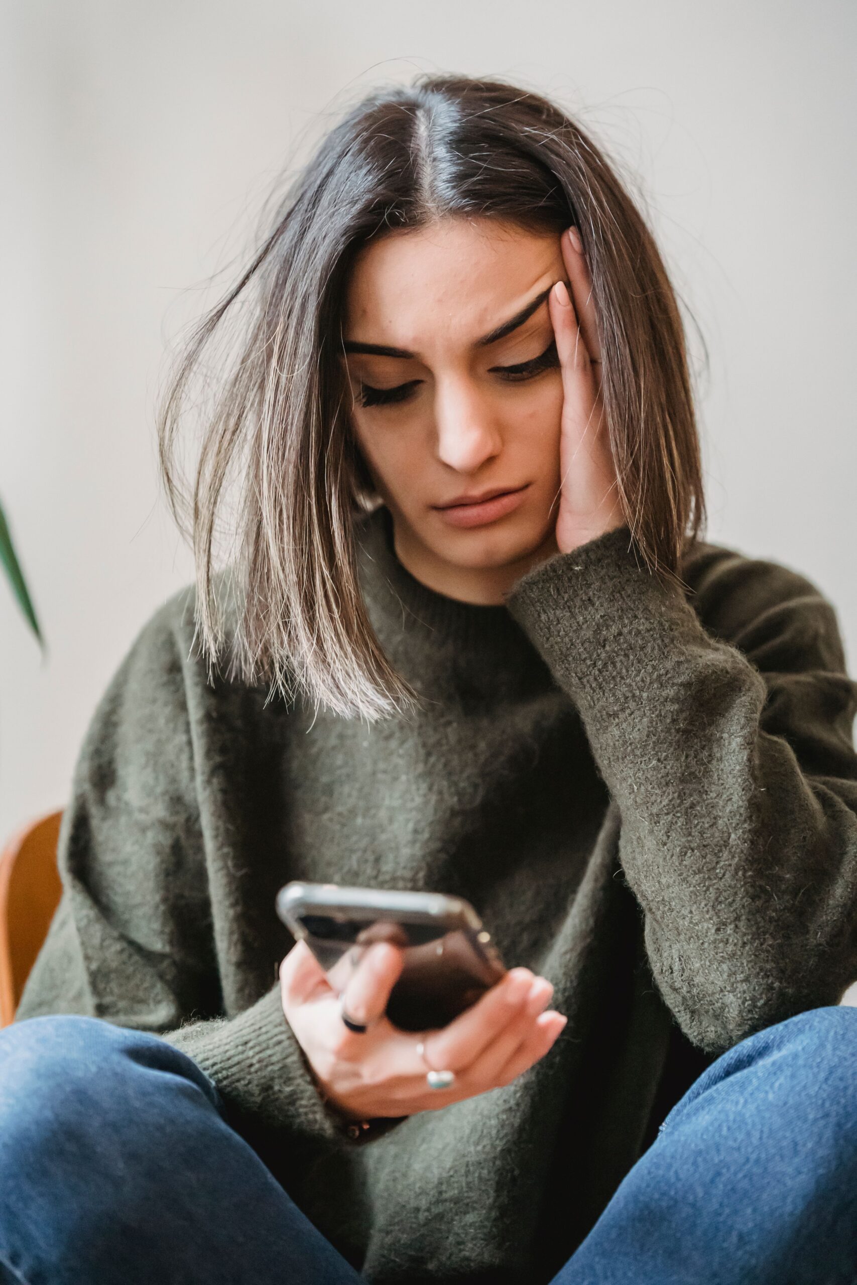 Jeune femme assise en tailleur, main sur le front, regardant son smartphone avec une expression d'inquiétude ou de stress