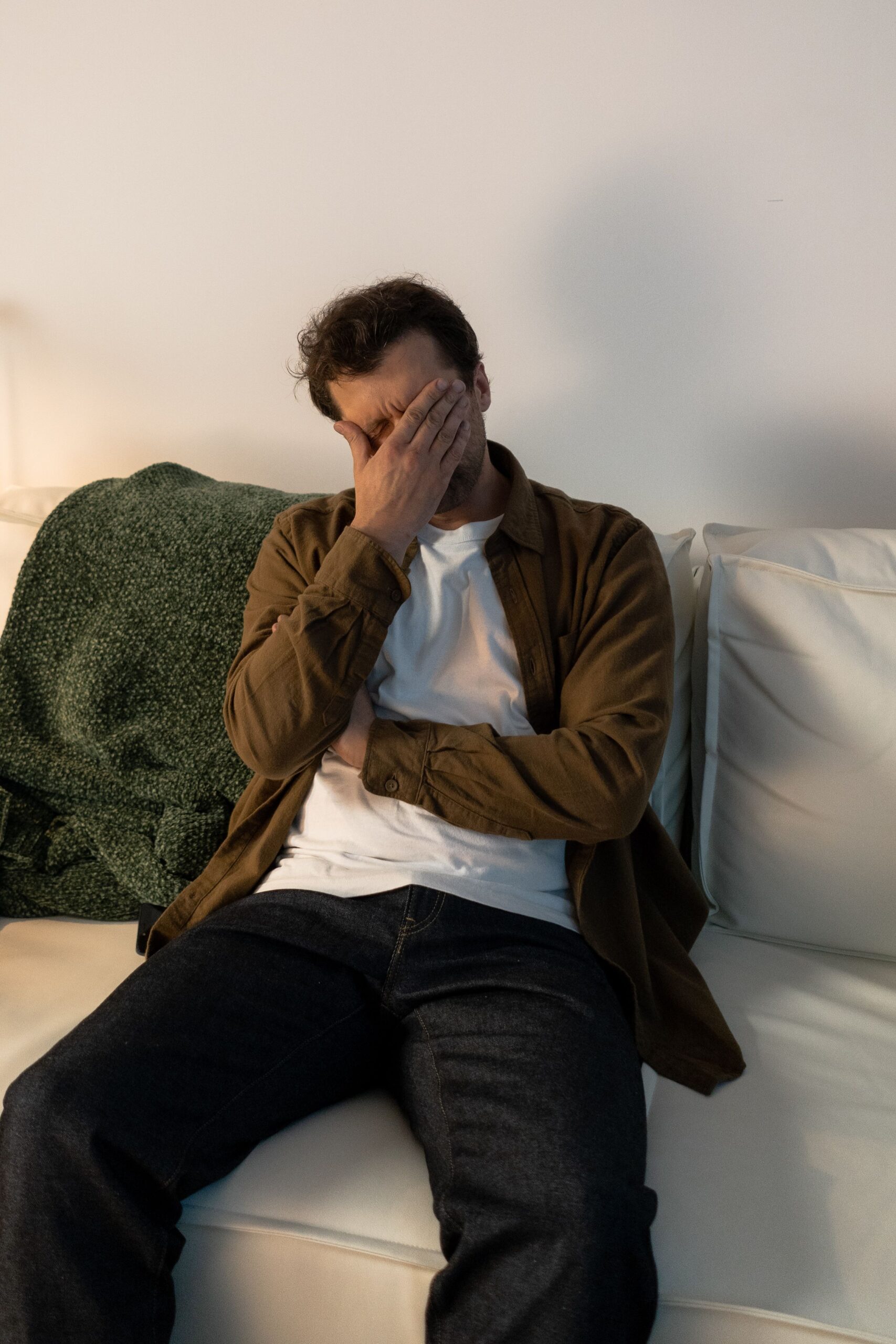 Homme assis sur un canapé blanc, se cachant le visage avec sa main, exprimant la fatigue, l'épuisement ou le désespoir